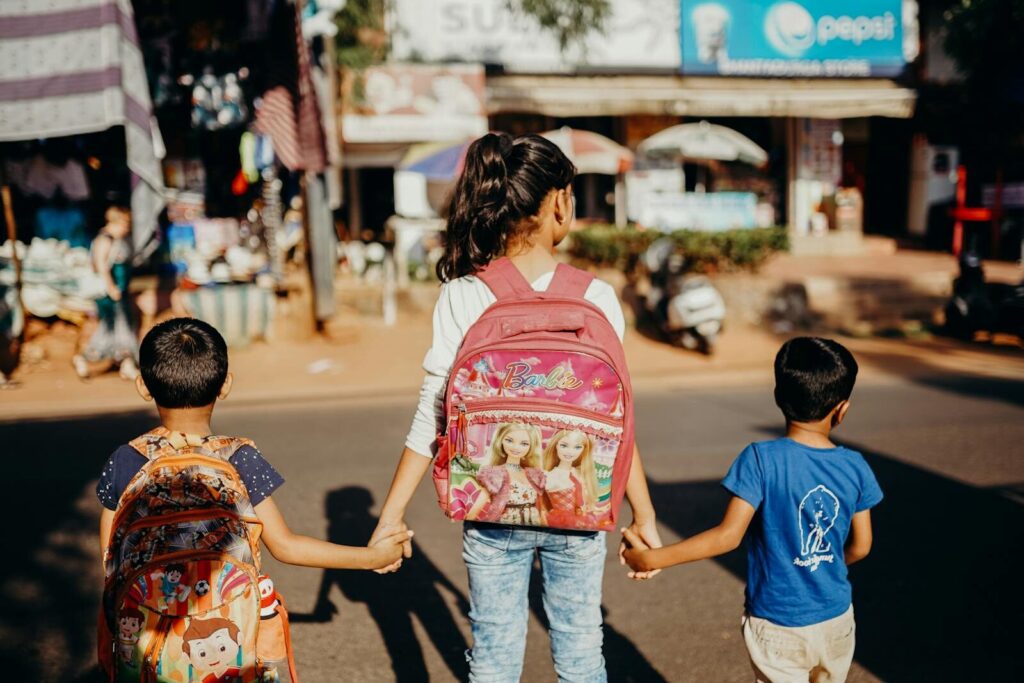 Three children with backpacks holding hands while crossing a street in India.